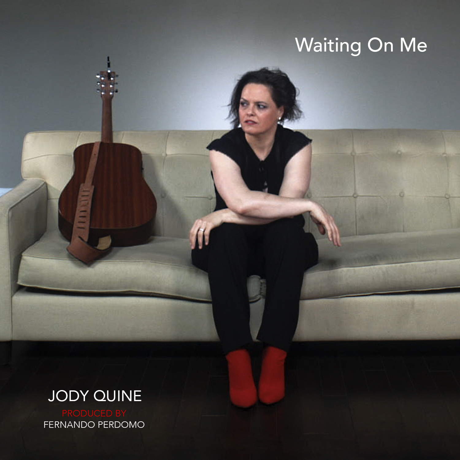 Jody Quine on a white couch with her guitar. She wears all black and is wearing red boots. The title of the song as well as Jody Quine and Produced by Fernando Perdomo are also in this visual.